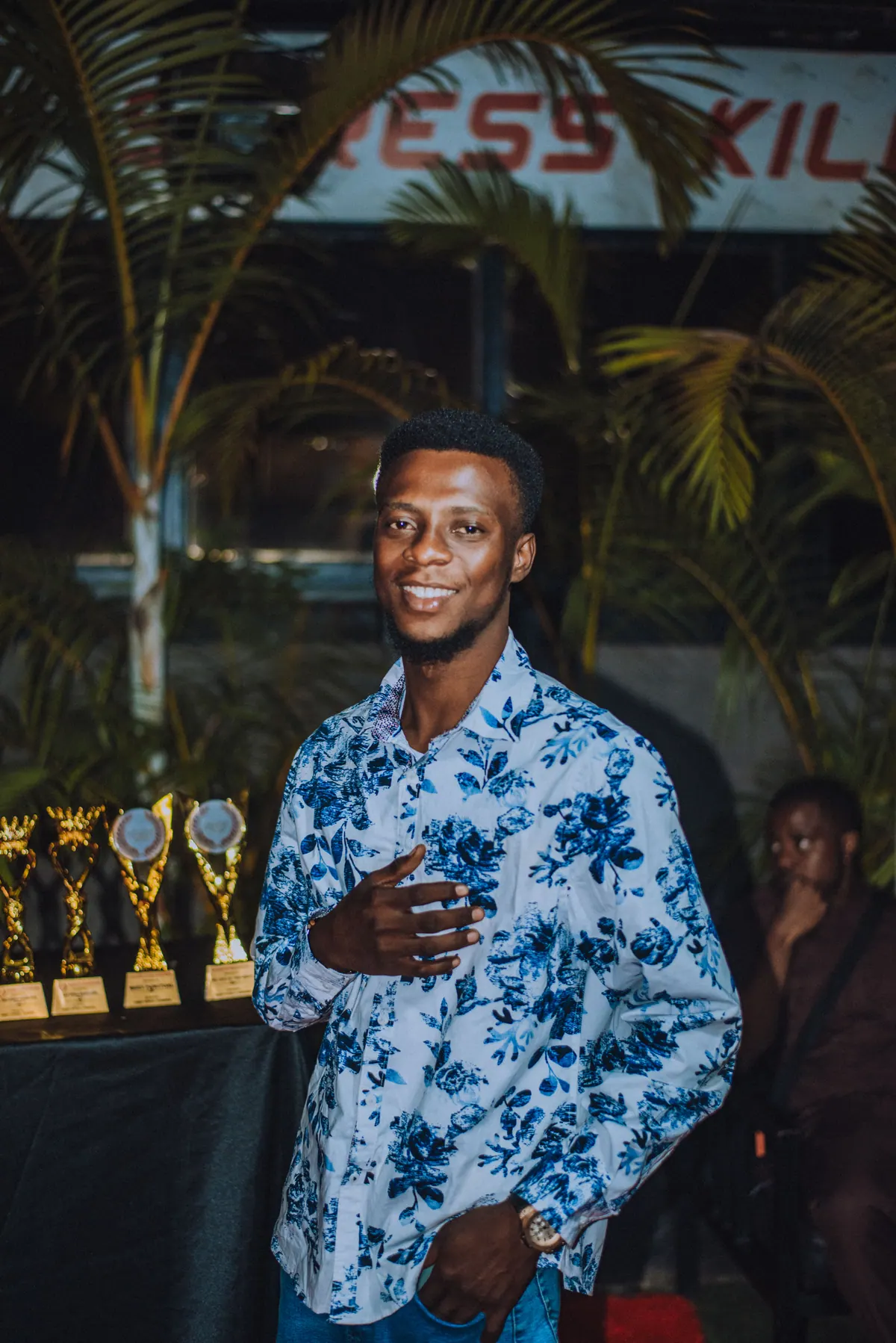 A man in a blue floral shirt smiles with a hand on his chest, trophies on the table behind him