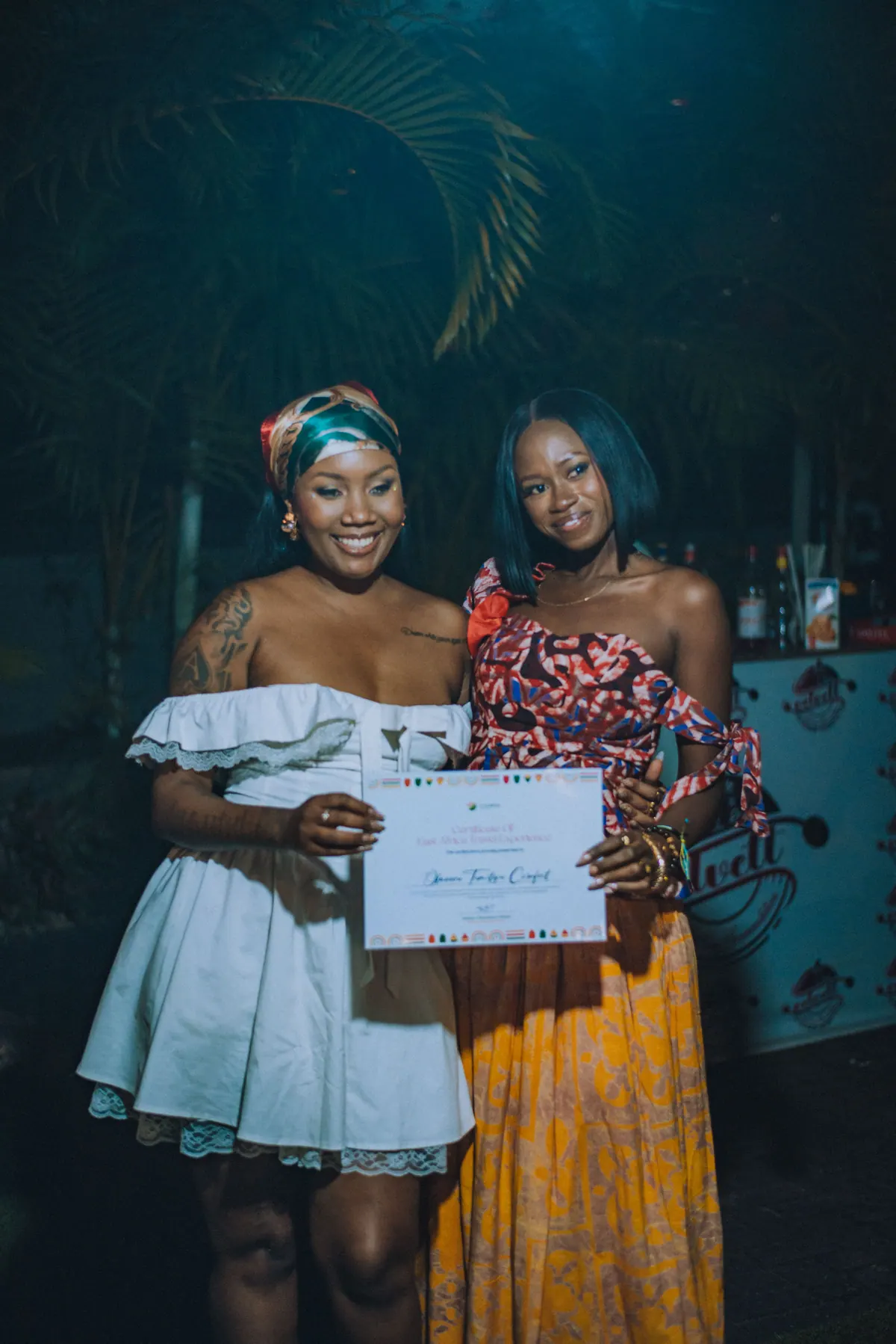 A woman in a print top and yellow skirt holds up her certificate beside the host in white