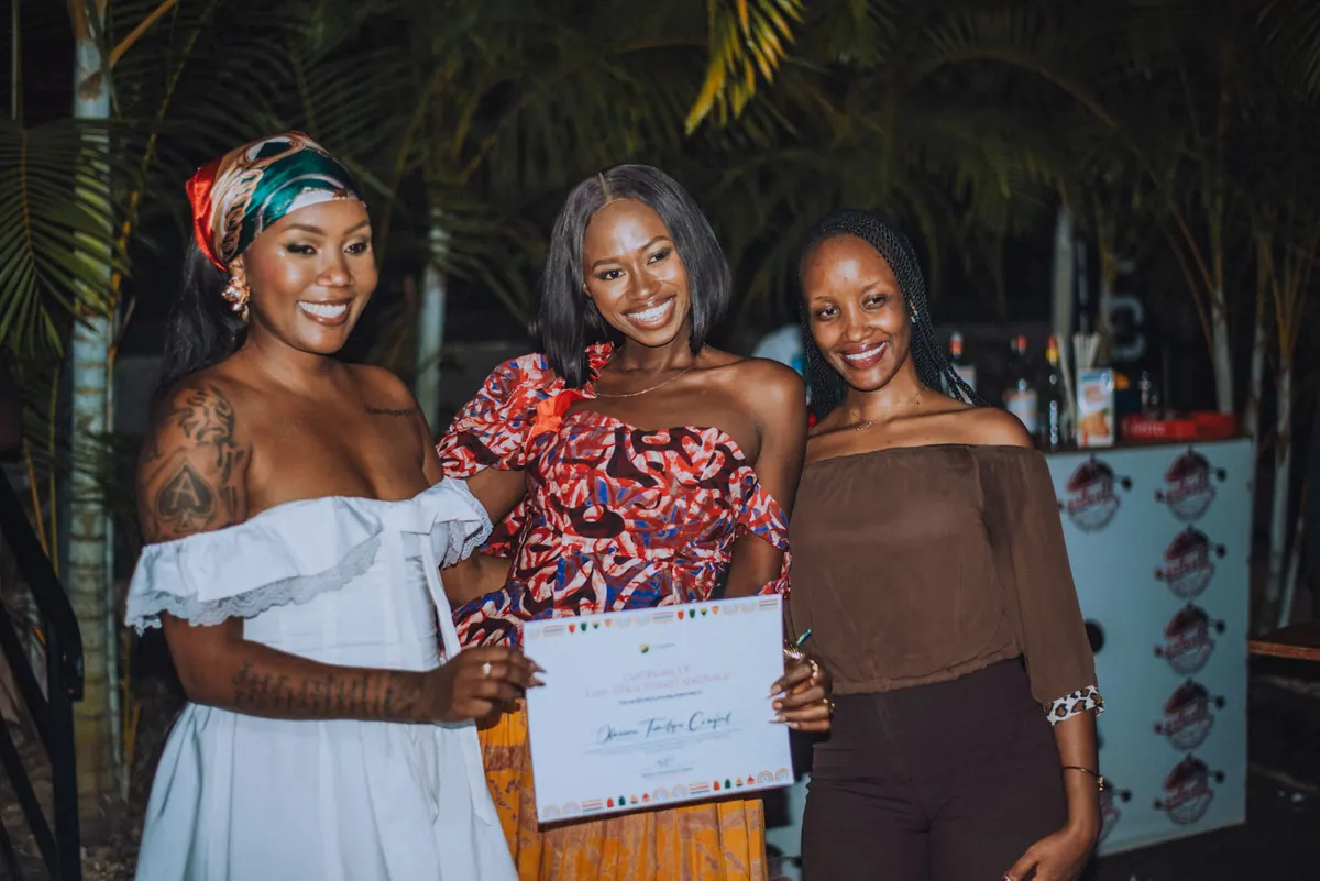 A woman in a red print top holds her certificate between the two hosts