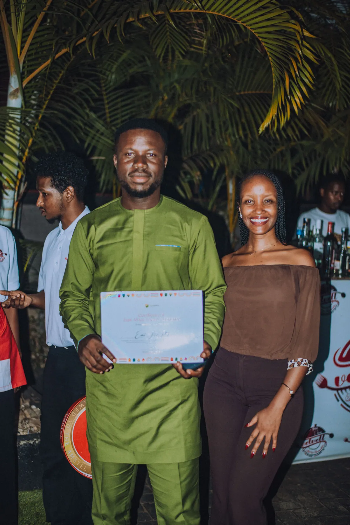 A man in a green kaftan holds his certificate beside a woman in a brown top