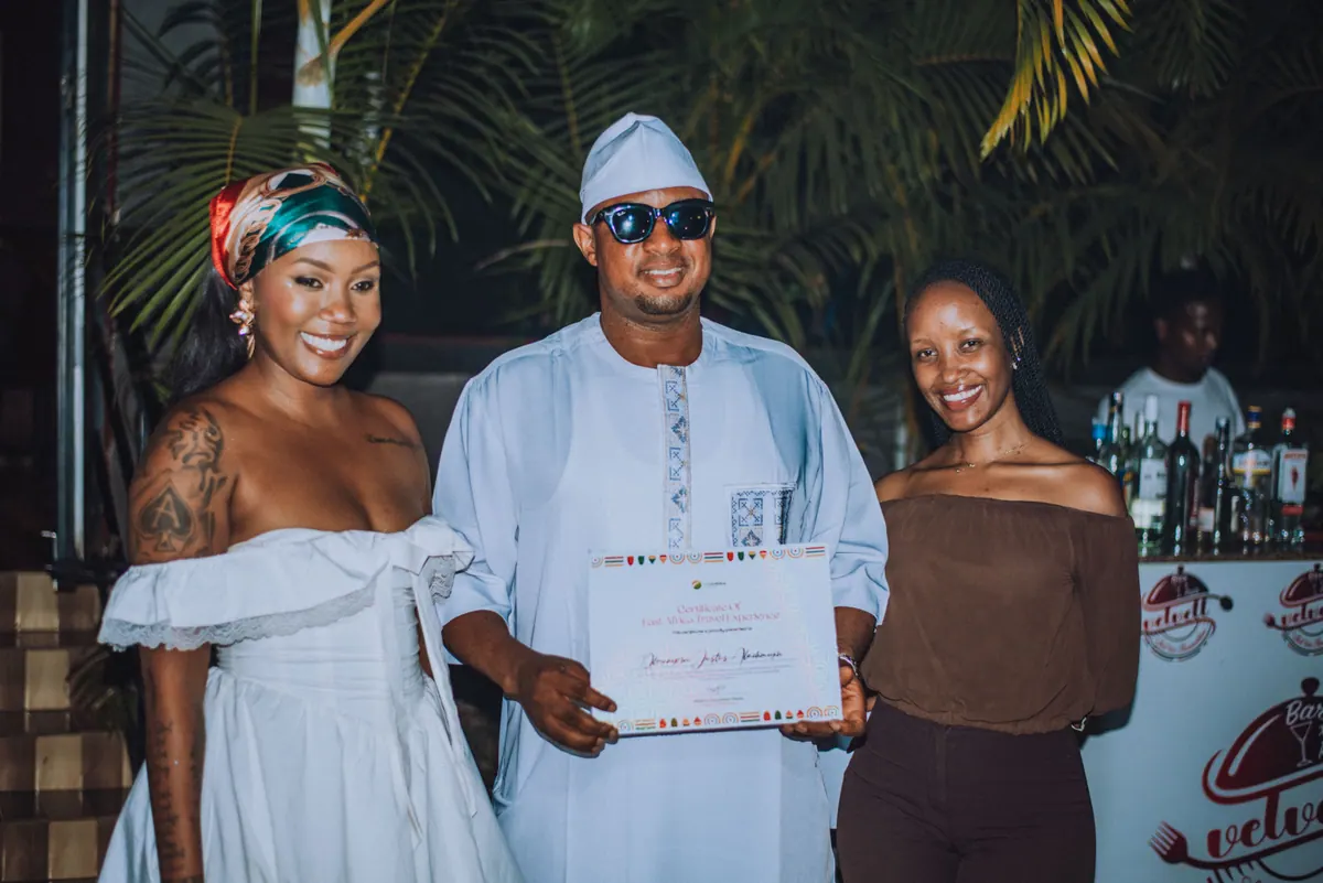A man in a white kaftan and cap holds a certificate between two women at the awards table