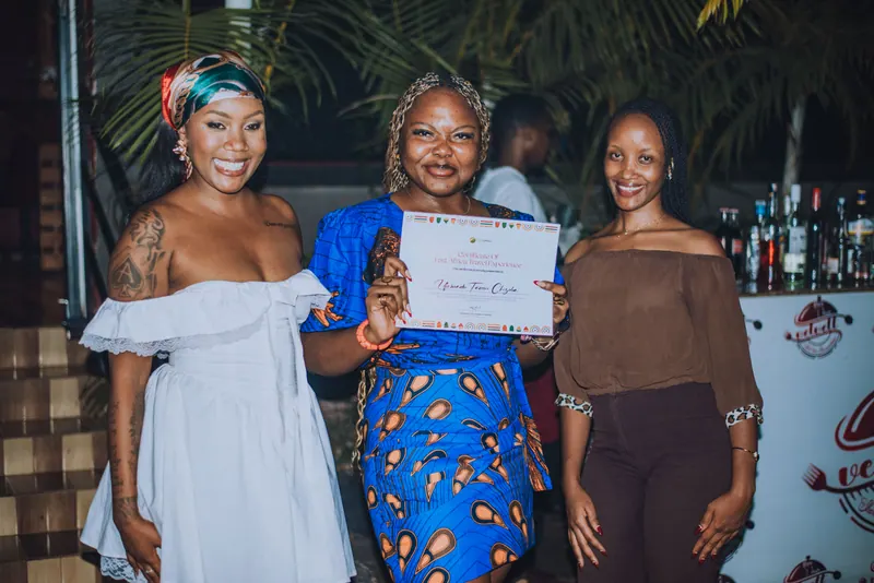 A woman in a blue ankara dress holds her certificate between the two hosts
