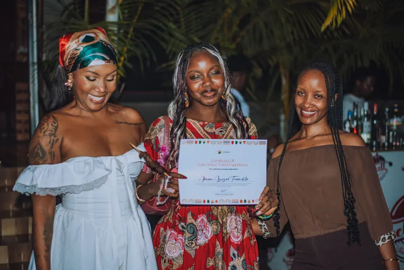 A woman in a red floral dress holds up her certificate between the two hosts