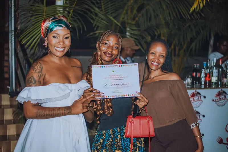 A woman in a teal dress with a red bag holds up her certificate