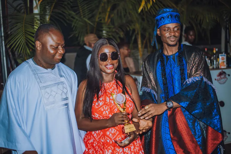 A woman in a red and white dress holds a gold trophy between two men in agbada