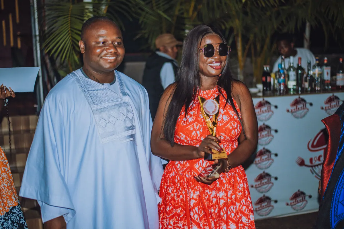 A woman in sunglasses and a red and white dress holds the Most Punctual trophy beside a man in a pale blue agbada