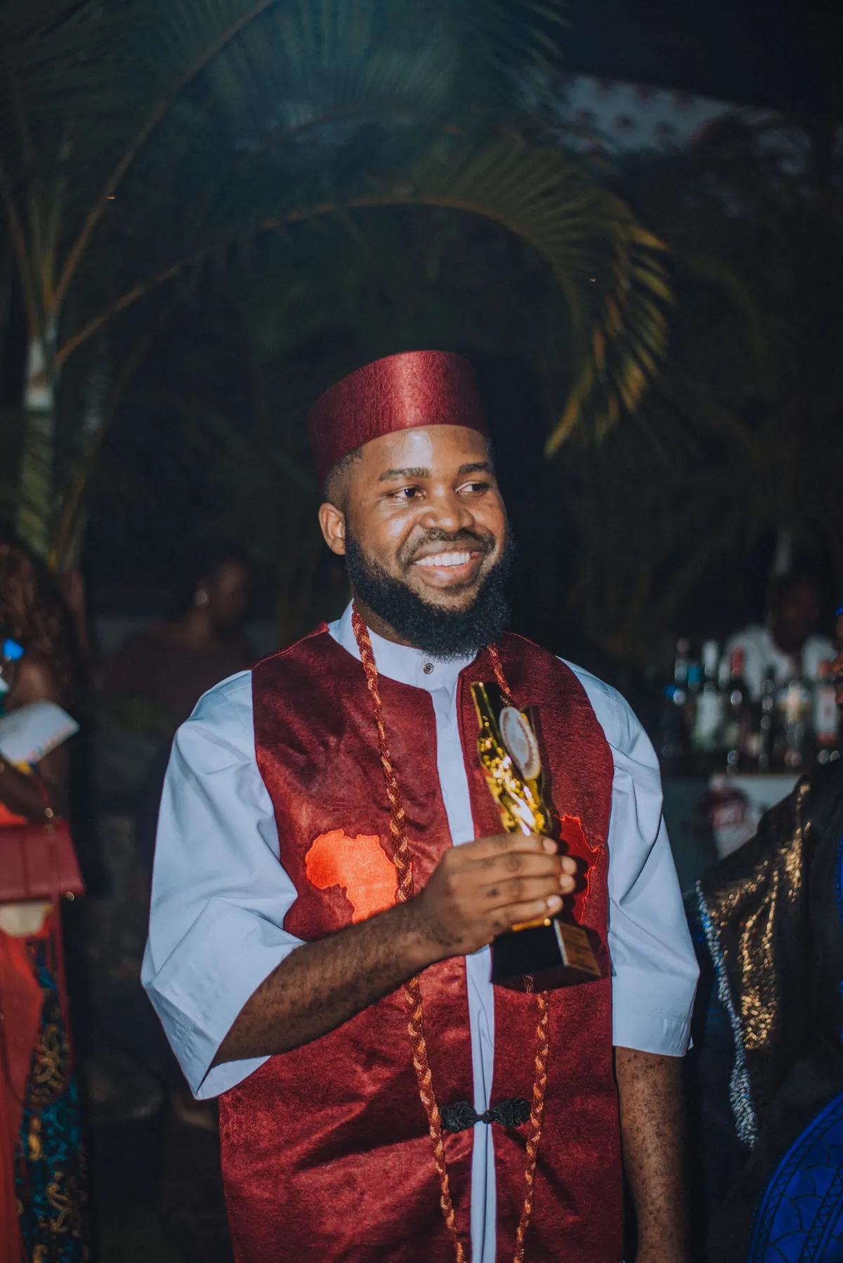 A bearded man in a red cap and red brocade waistcoat smiles, holding a gold trophy