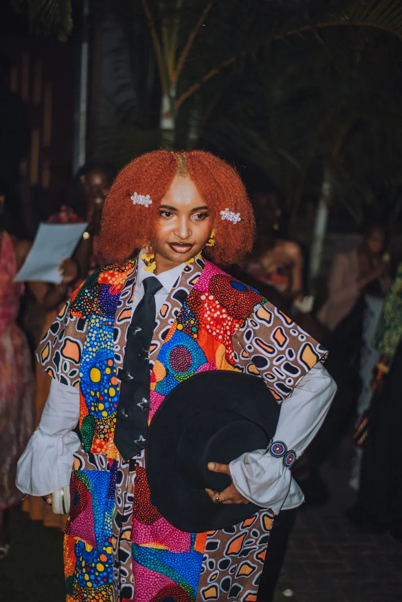 A woman with a red afro in a patchwork ankara jacket, white shirt and black tie holds a black hat on her hip