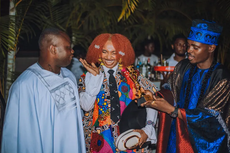 A woman with a red afro and a patchwork jacket takes a bow as she receives a trophy from the hosts