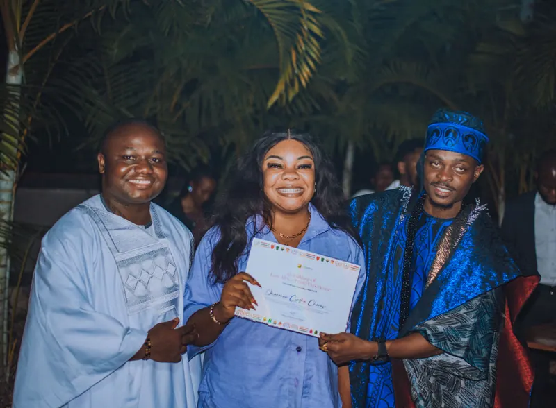 A woman in a pale blue shirt dress holds her certificate between the two men presenting