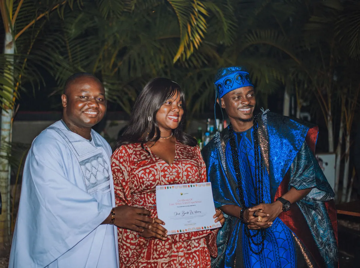 A woman in a red and white adire dress holds her certificate between the two men presenting