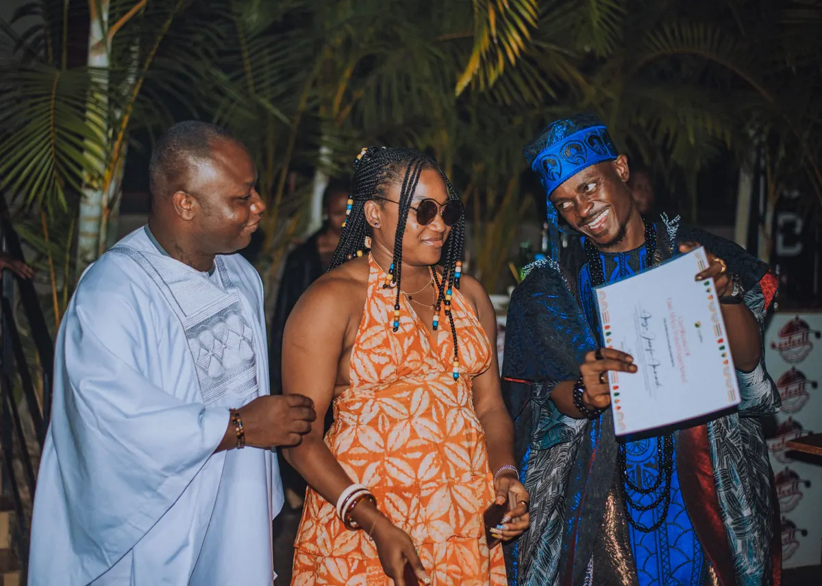 A woman in sunglasses and an orange print dress stands between the hosts as her certificate is read out