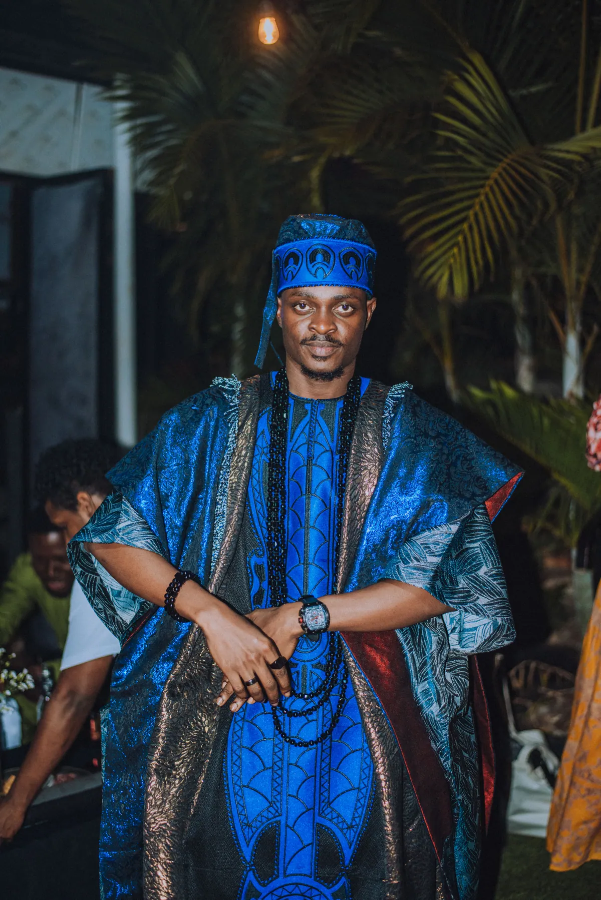 A man in a blue aso-oke cap and layered blue agbada with long black beads stands with his hands clasped
