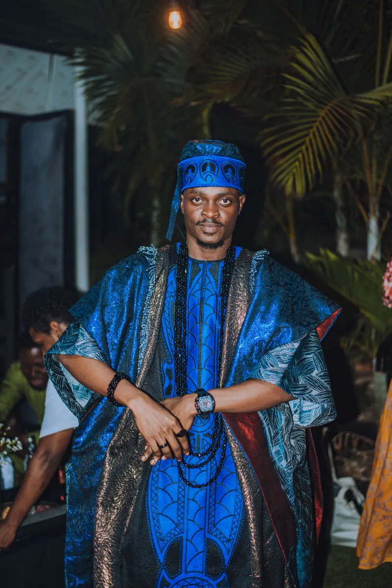 A man in a blue aso-oke cap and layered blue agbada with long black beads stands with his hands clasped