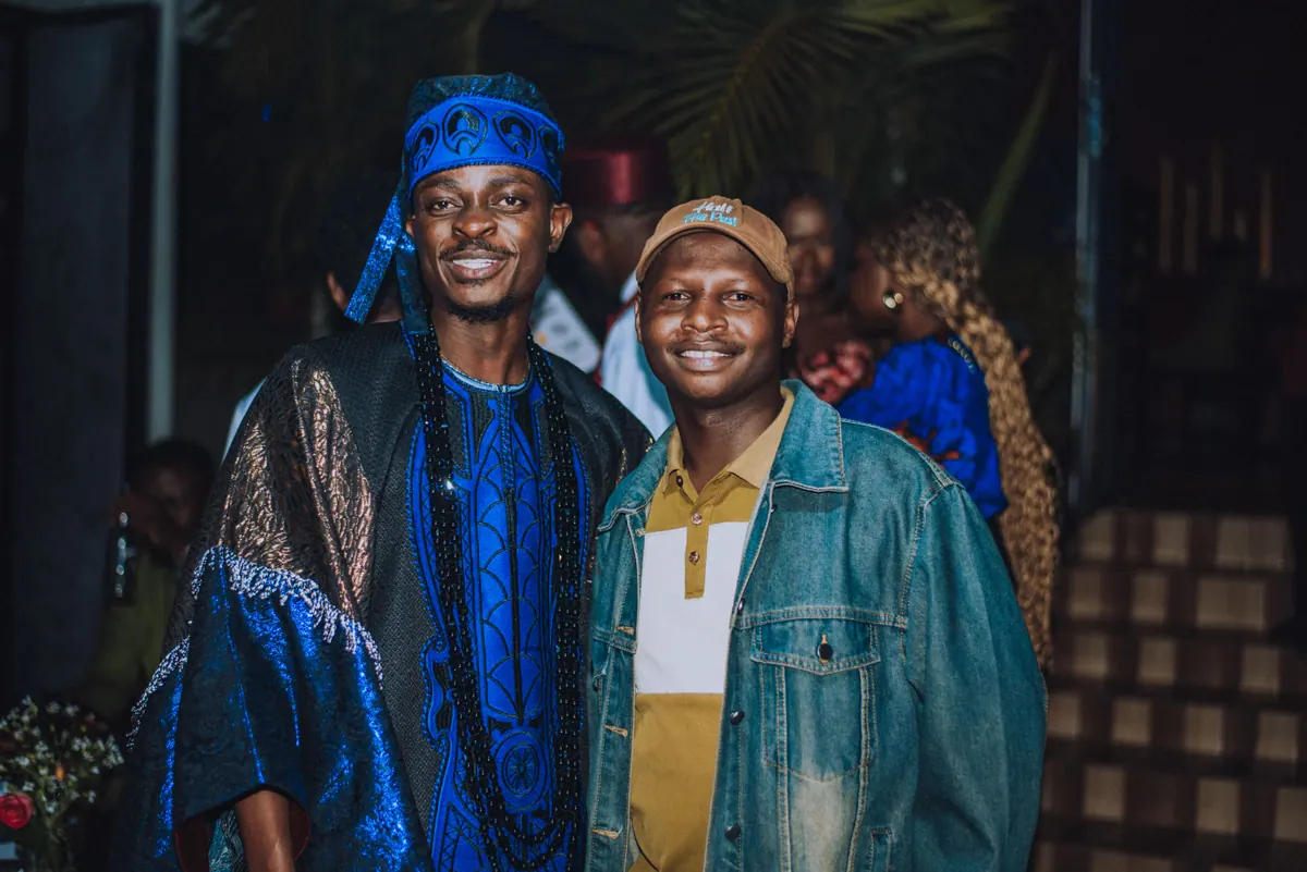 A man in a blue agbada and a man in a denim jacket and brown cap smile side by side