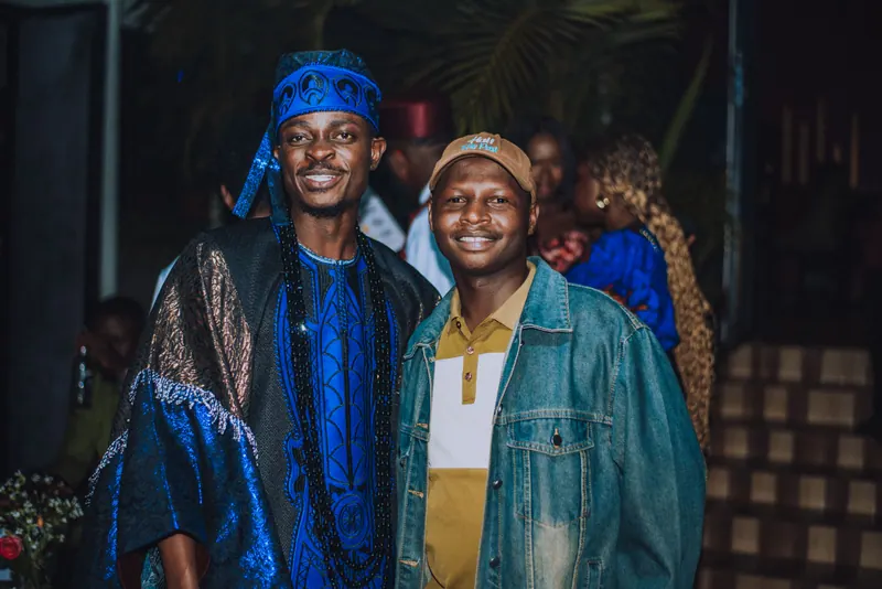 A man in a blue agbada and a man in a denim jacket and brown cap smile side by side