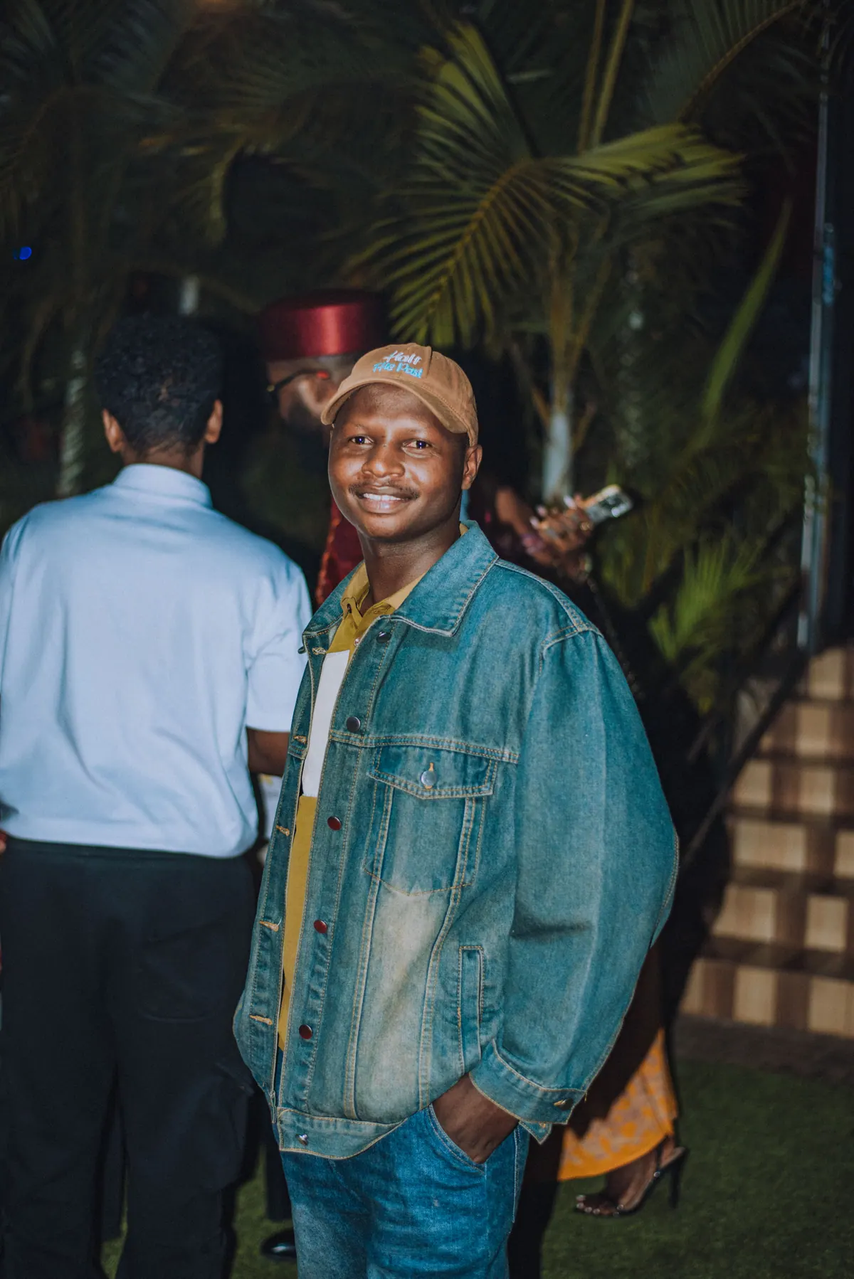 A man in a brown cap and an oversized denim jacket smiles on the lawn