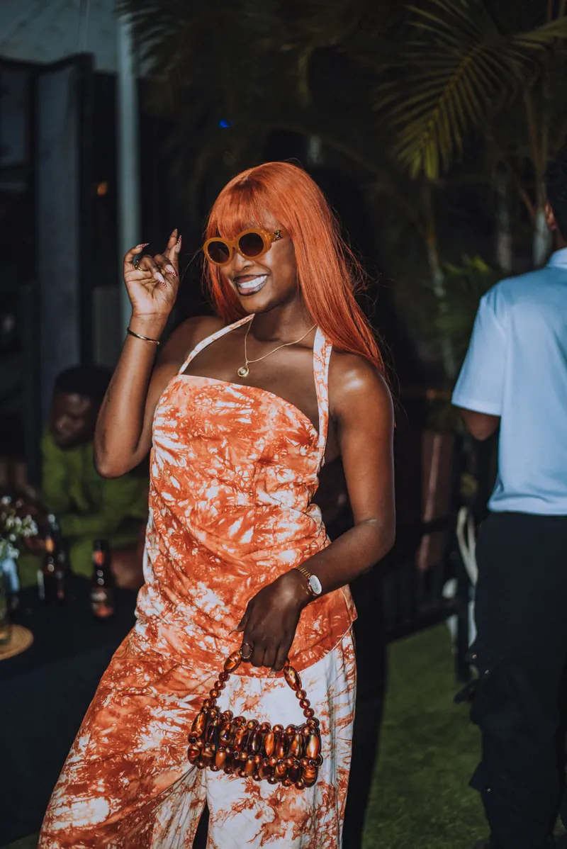 A woman with a copper fringe wig, round sunglasses and an orange tie-dye set dances holding a beaded bag