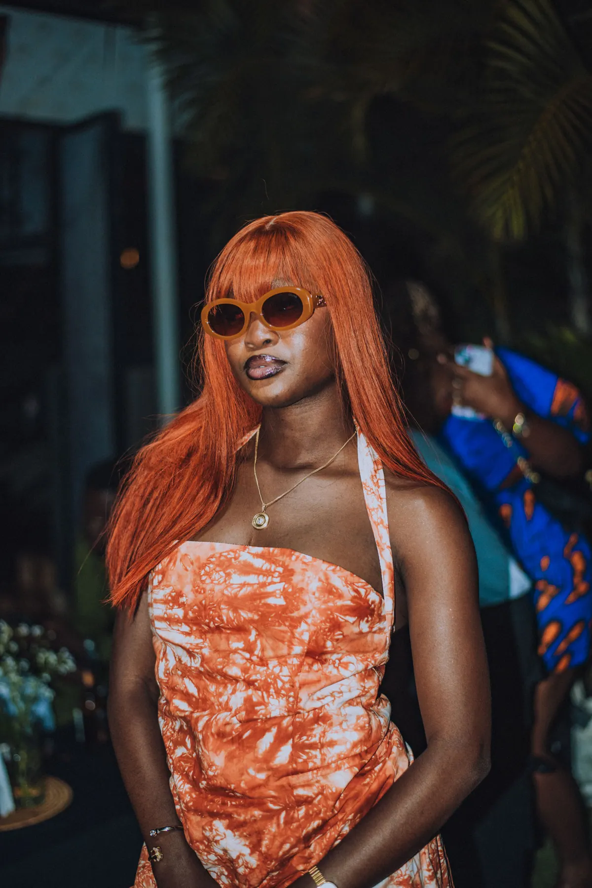 A woman with long copper hair and round sunglasses poses in an orange tie-dye halter set