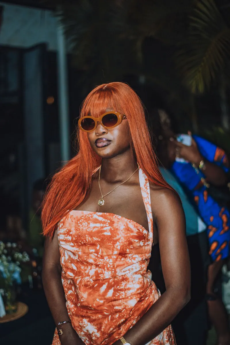 A woman with long copper hair and round sunglasses poses in an orange tie-dye halter set