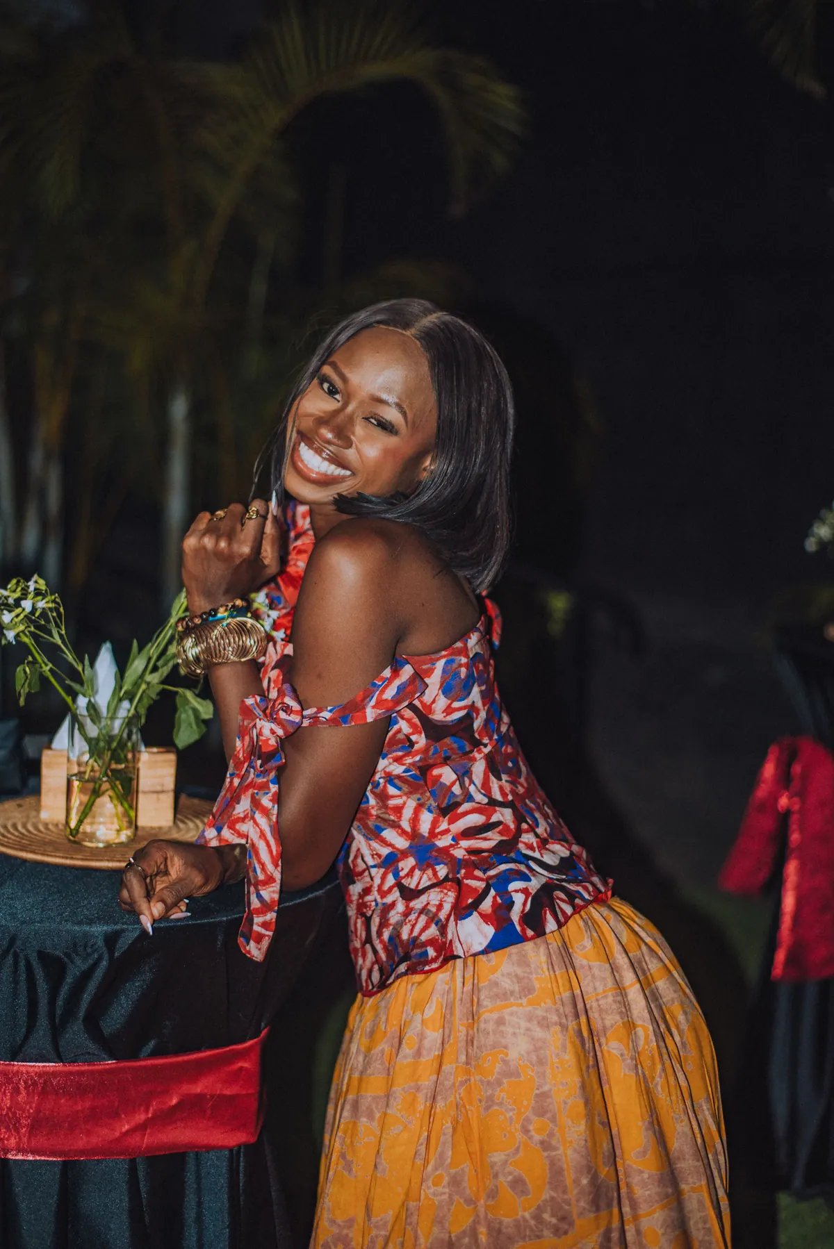 A woman in a red and blue print top and yellow skirt leans on a table, smiling over her shoulder
