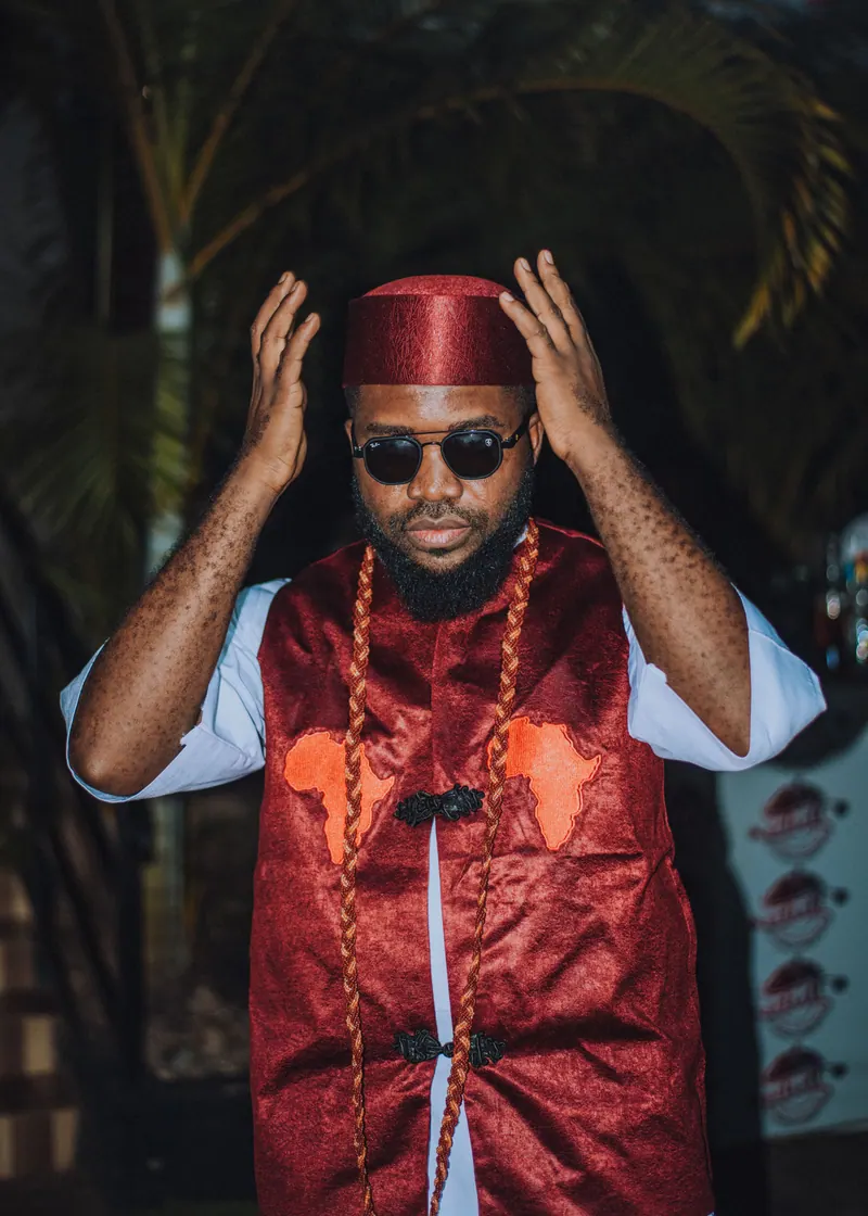 A man in a red cap, sunglasses and a red brocade waistcoat with coral beads raises both hands to his cap