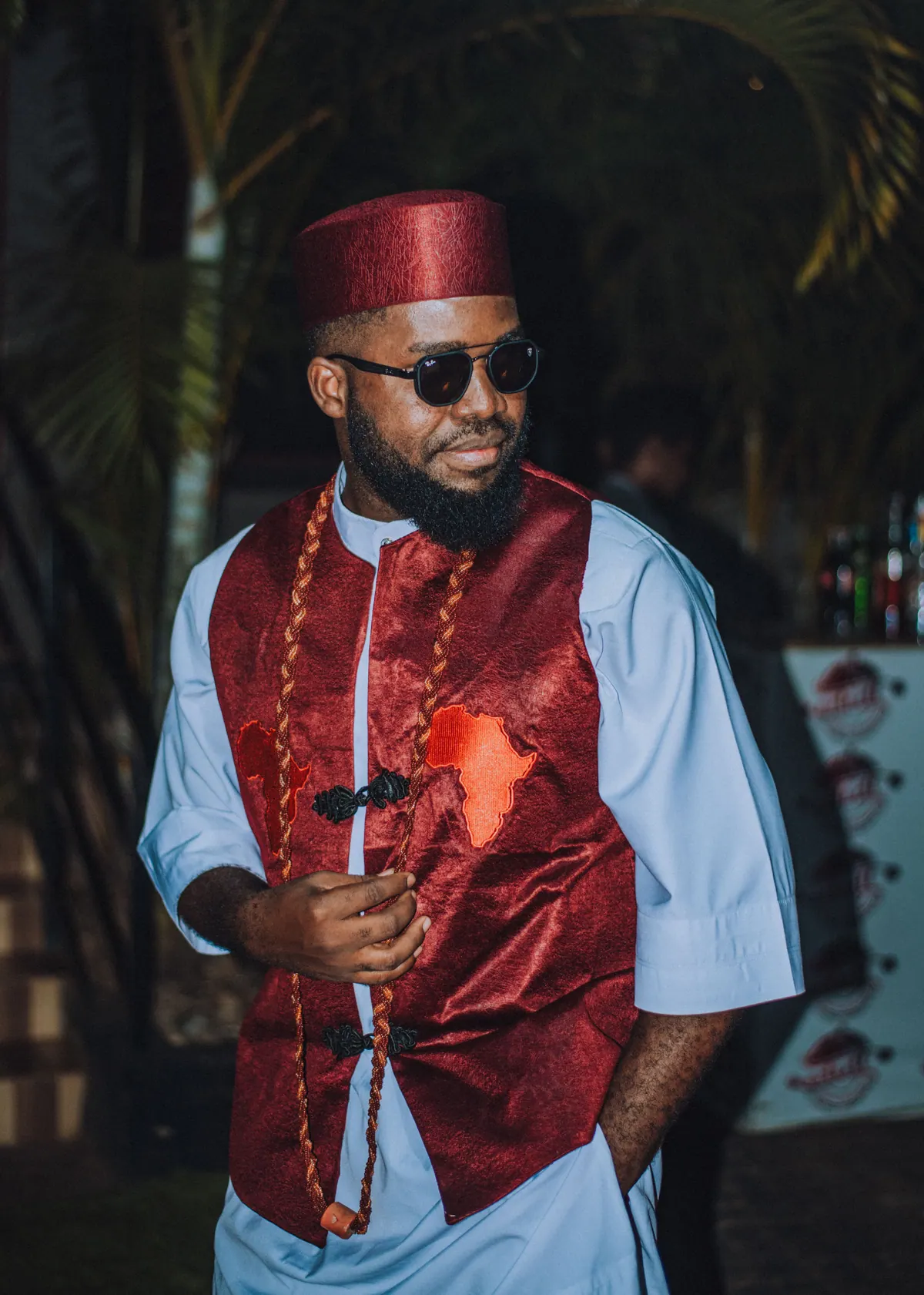 A man in a red cap and red waistcoat with coral beads looks past the camera