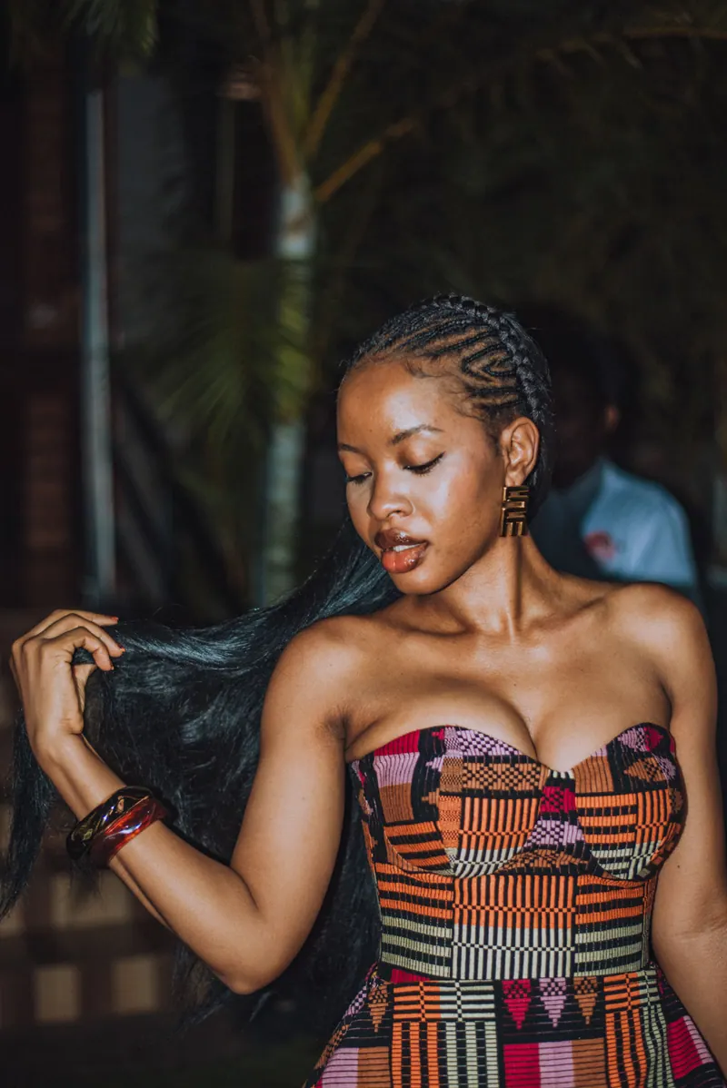 A woman with cornrows and gold earrings in a strapless kente-pattern dress sweeps her hair to one side