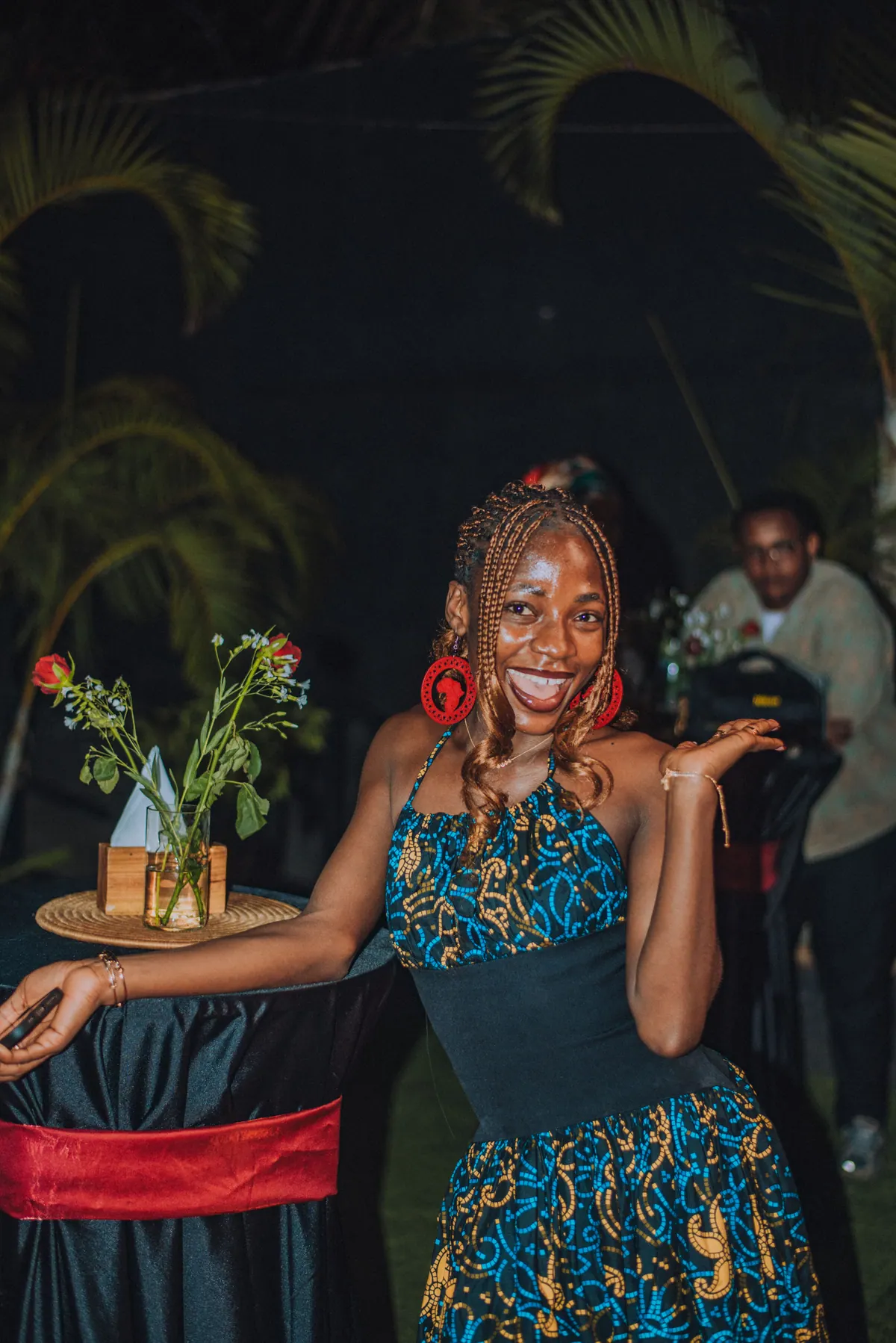 A woman in red beaded earrings and a blue ankara dress dances with open hands