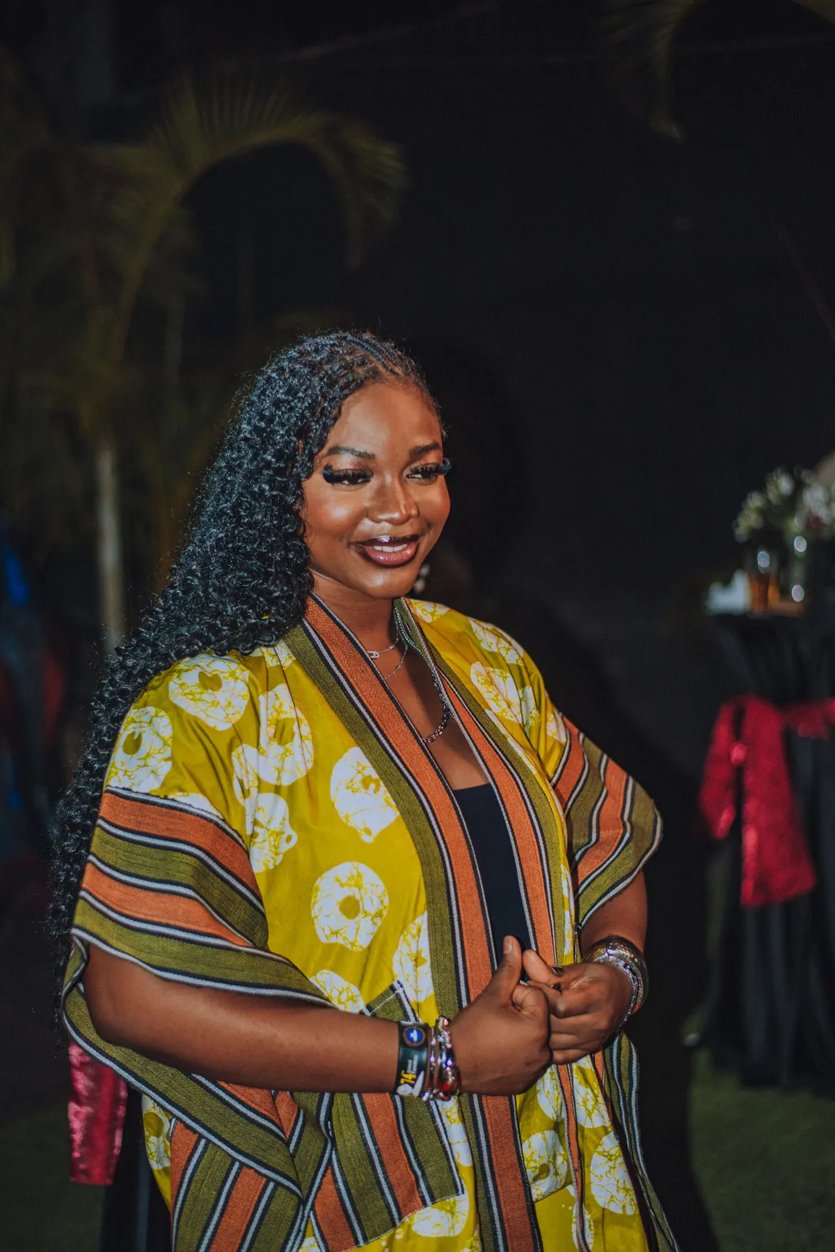 A woman with long curls in a yellow adire kimono smiles with her hands together
