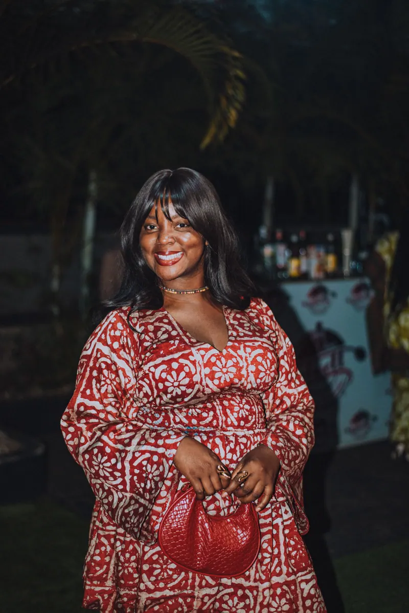 A woman in a red and white adire dress smiles, holding a red woven bag