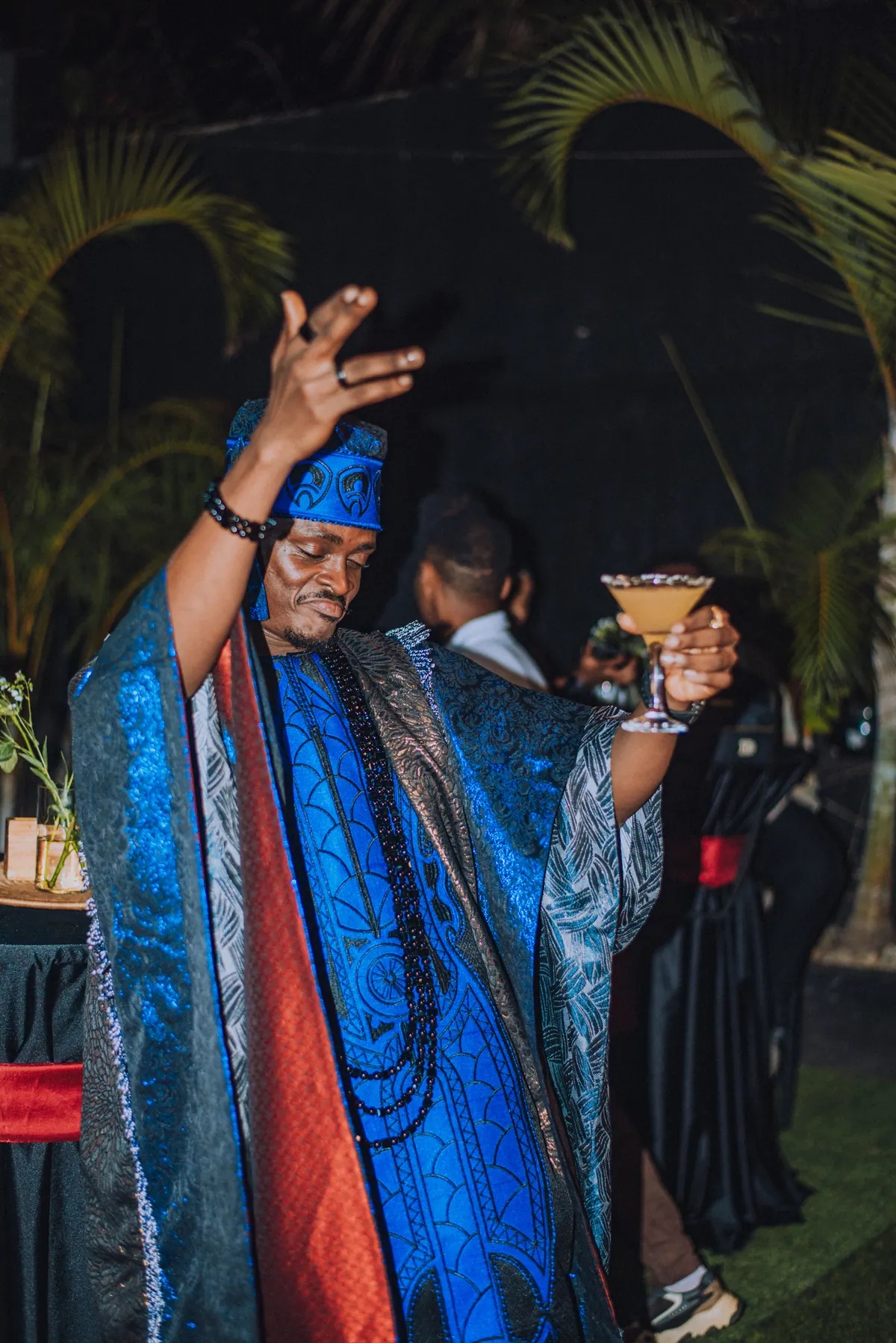 A man in a blue agbada raises a cocktail glass and one hand in the air