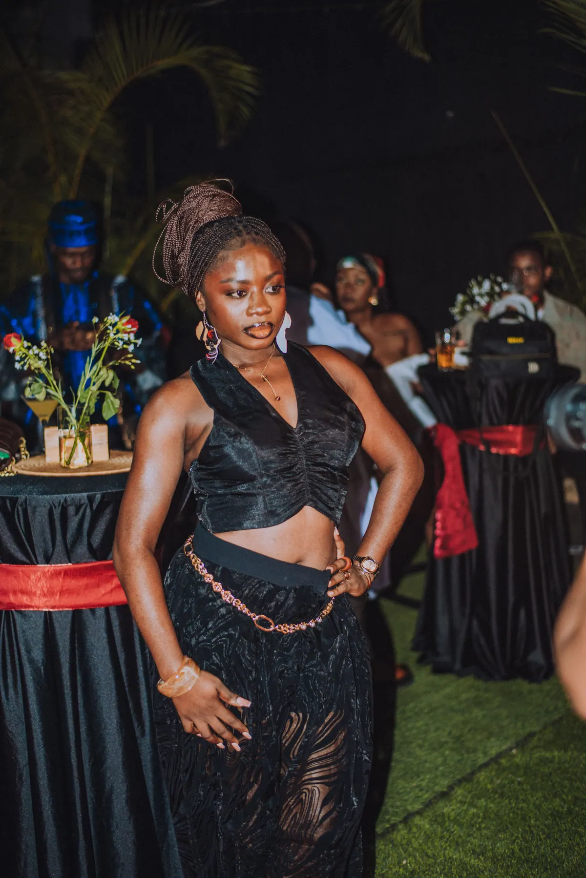 A woman in a black satin top and lace skirt with waist beads stands by the cocktail tables