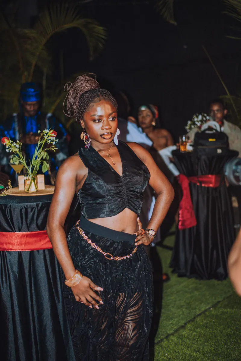 A woman in a black satin top and lace skirt with waist beads stands by the cocktail tables