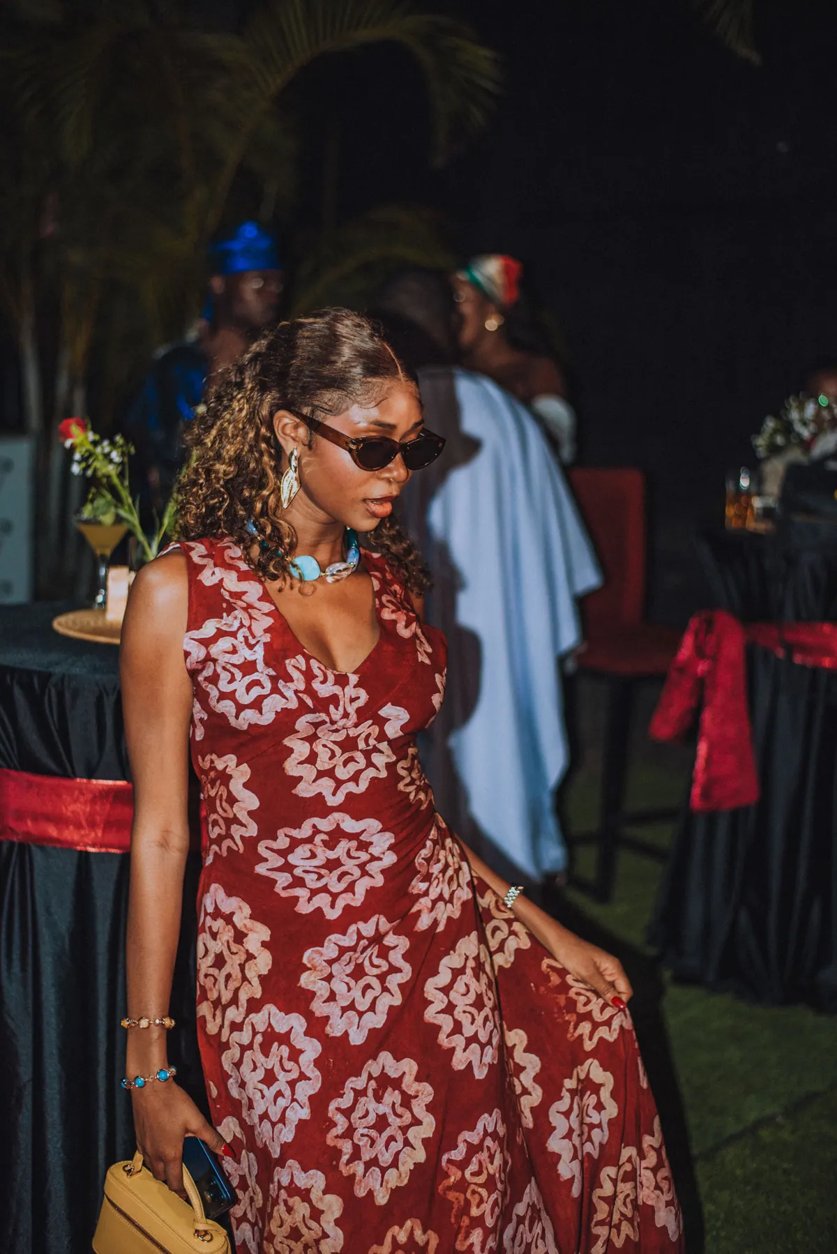 A woman in sunglasses and a red and white print dress walks across the lawn carrying a yellow bag