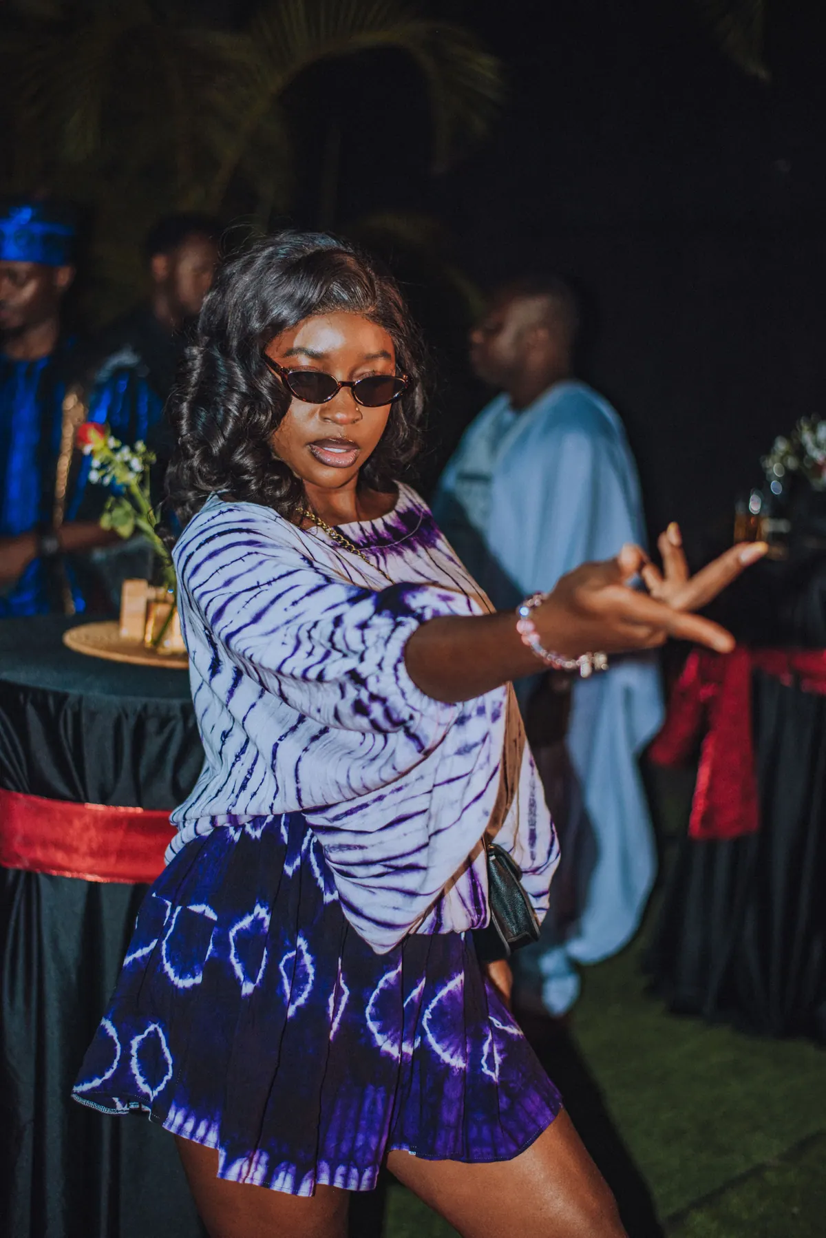 A woman in sunglasses and a purple tie-dye set points across the lawn mid-dance