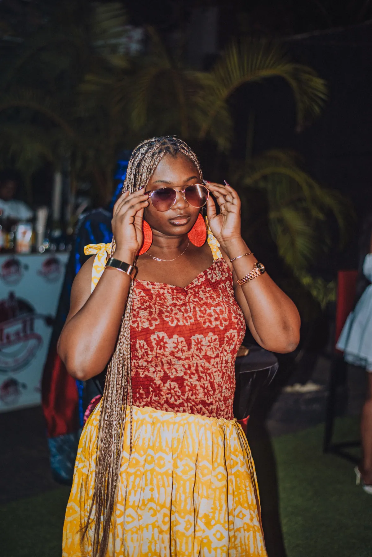 A woman with long braids and big red earrings adjusts her sunglasses, wearing a red top and yellow skirt