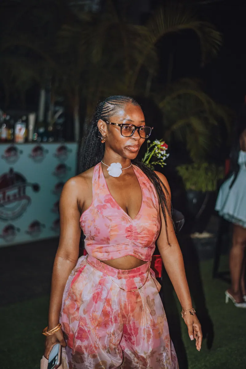 A woman in glasses and a pink floral halter set walks through the party holding her phone