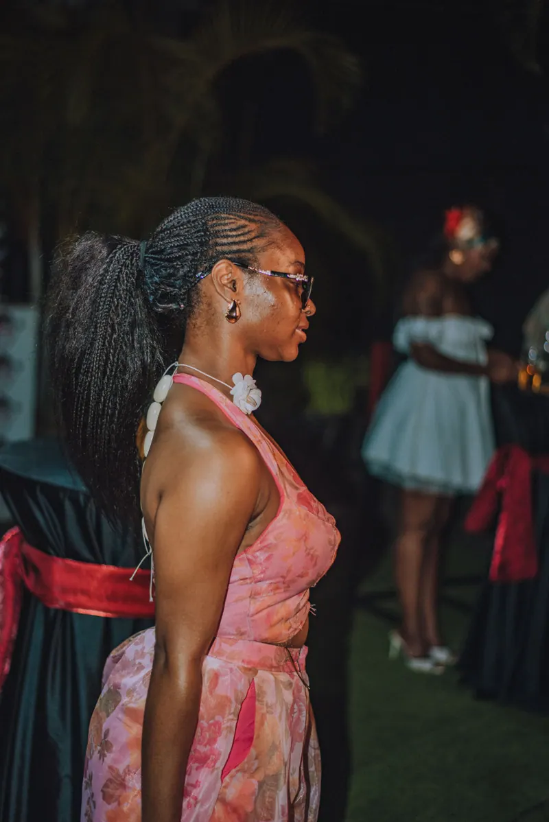 A woman in a pink halter set in profile, her long ponytail threaded with cowrie shells