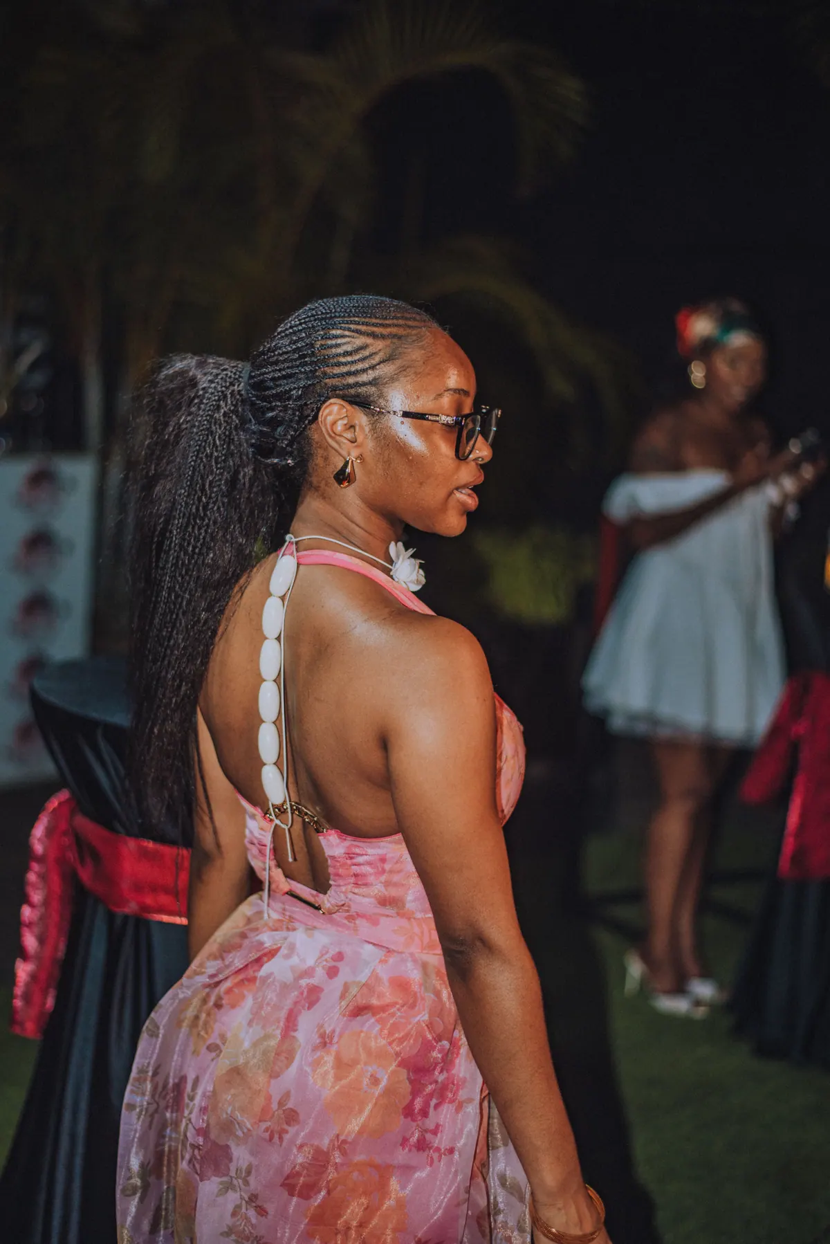 A woman in a pink halter set looks back over her shoulder, cowrie shells running down her ponytail
