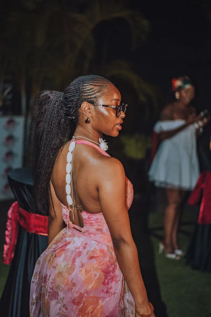 A woman in a pink halter set looks back over her shoulder, cowrie shells running down her ponytail