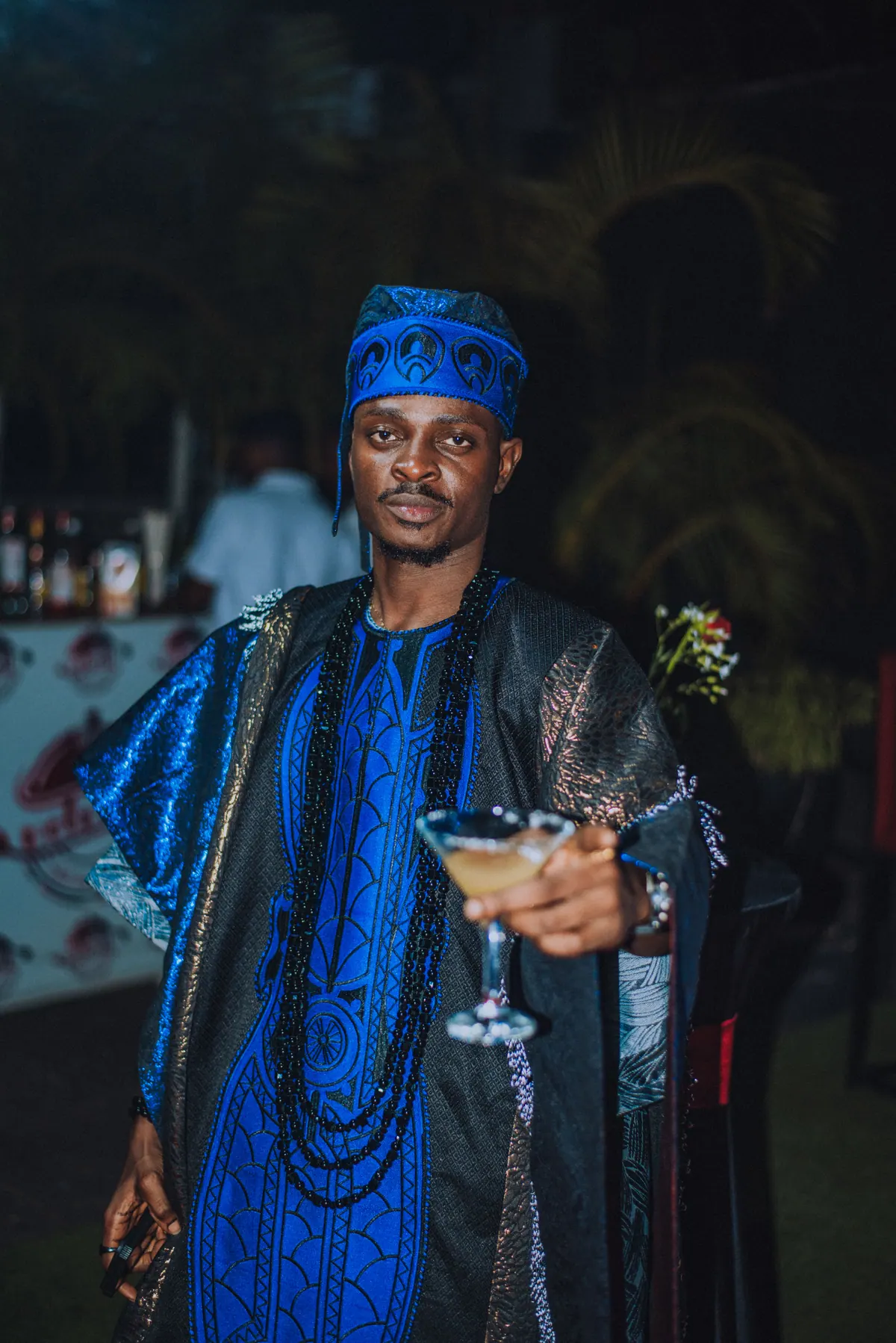 A man in a blue aso-oke cap and blue agbada with long beads holds a cocktail out to the camera