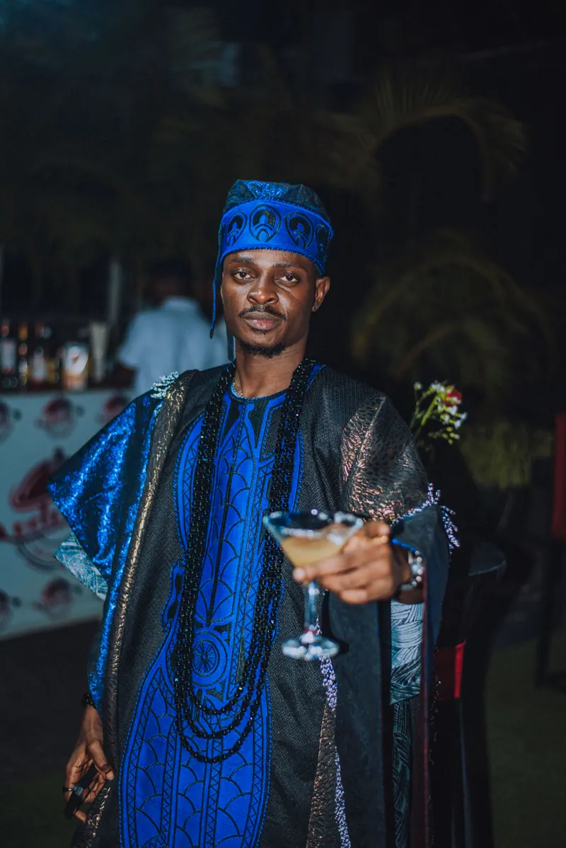 A man in a blue aso-oke cap and blue agbada with long beads holds a cocktail out to the camera