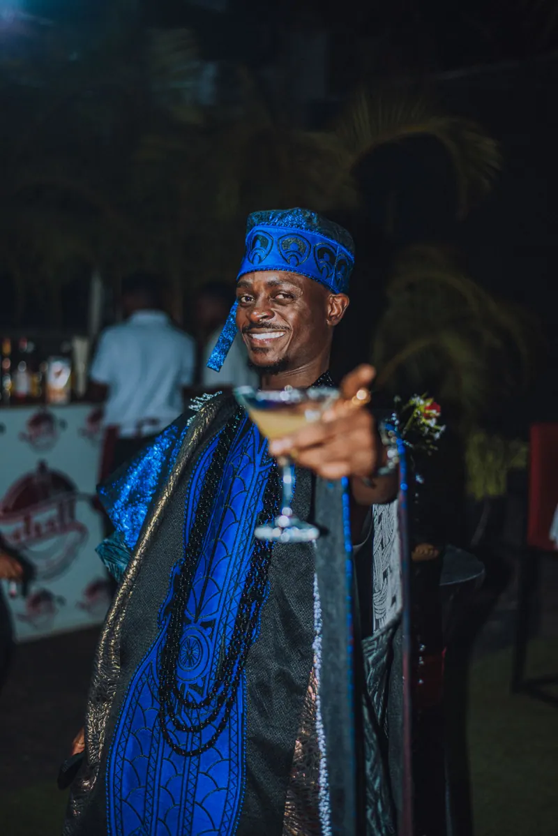 A man in a blue agbada laughs and holds a cocktail glass out to the camera