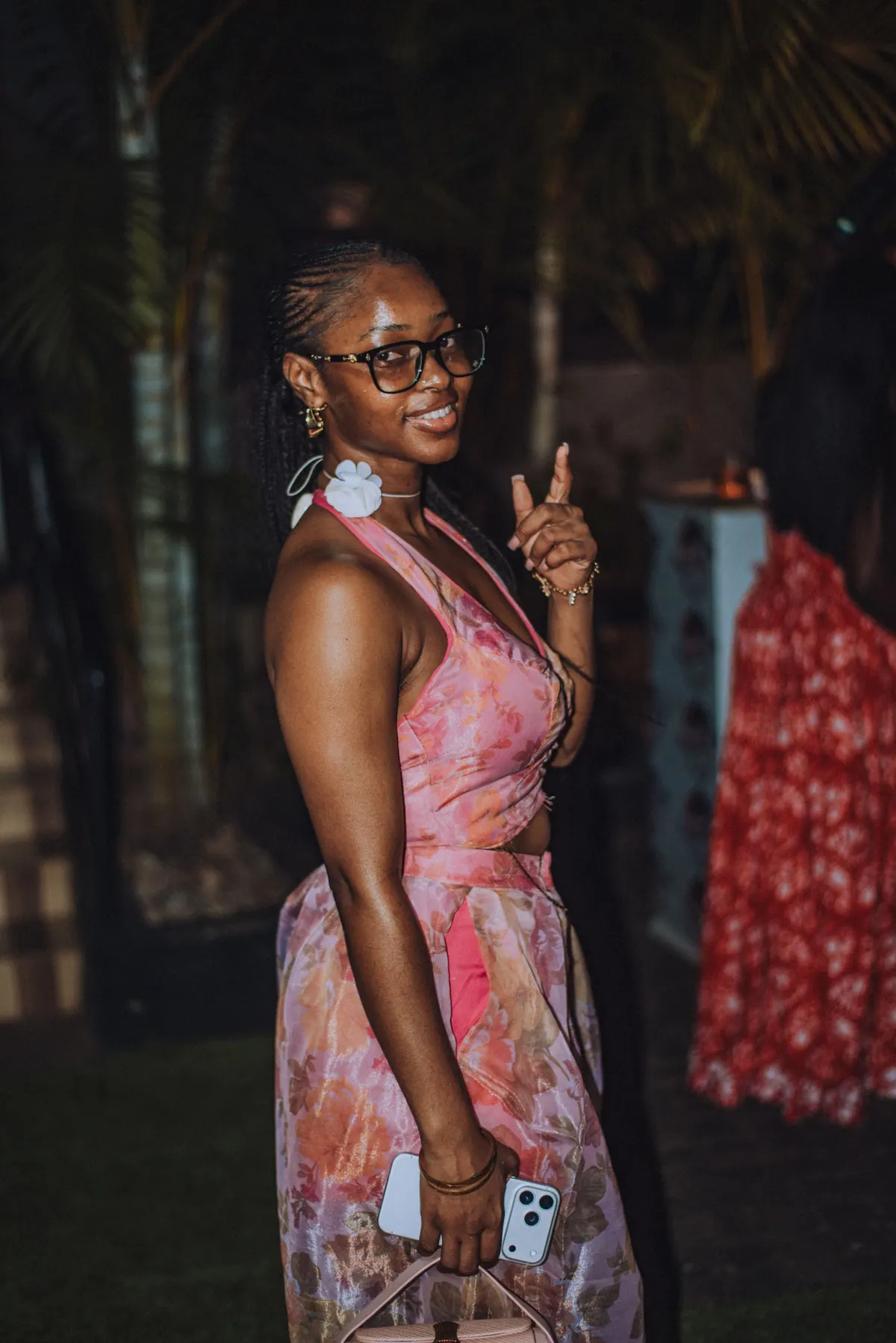 A woman in glasses and a pink floral dress smiles and points over her shoulder