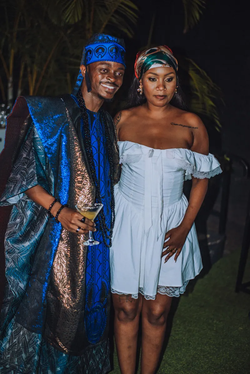 A man in a blue agbada and a woman in a white off-shoulder dress and headwrap stand together
