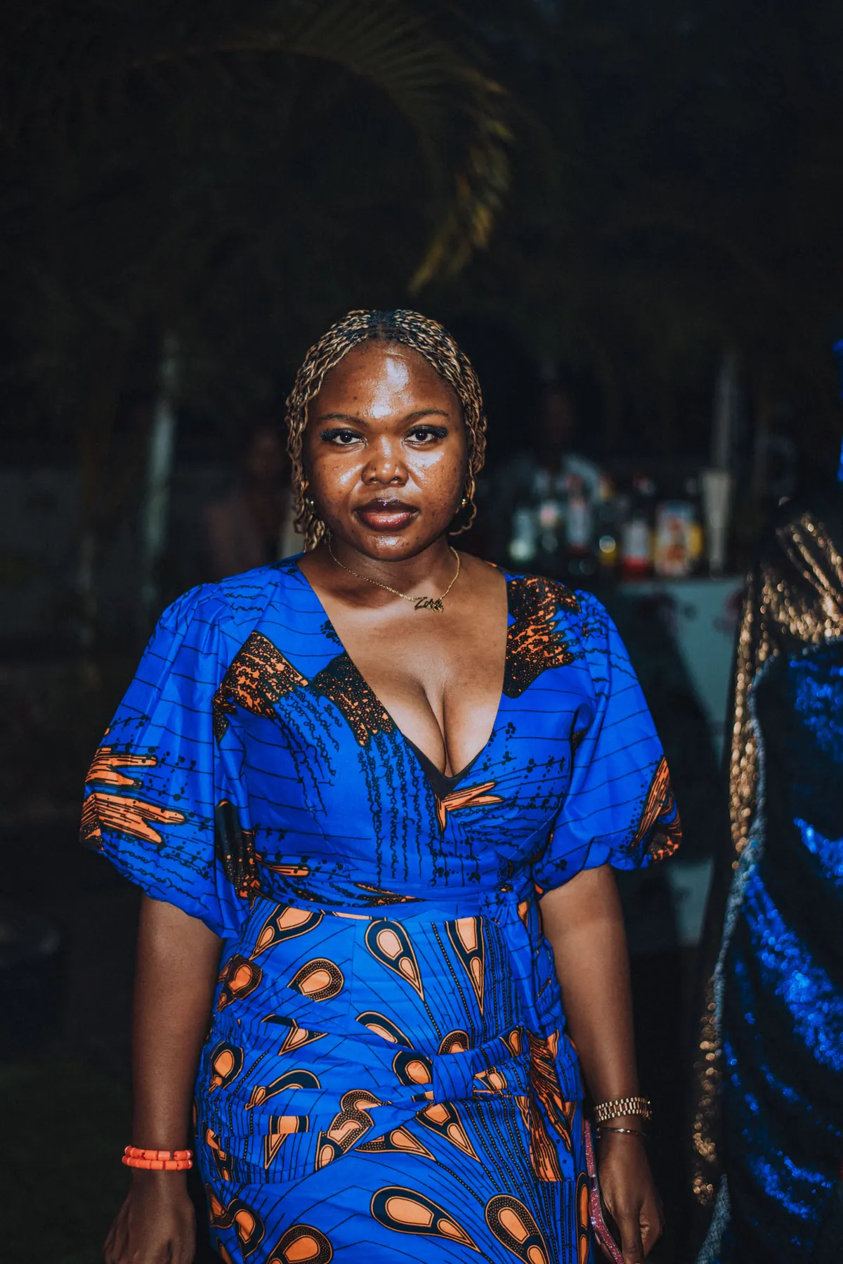 A woman with braided hair in a bright blue ankara dress looks straight at the camera