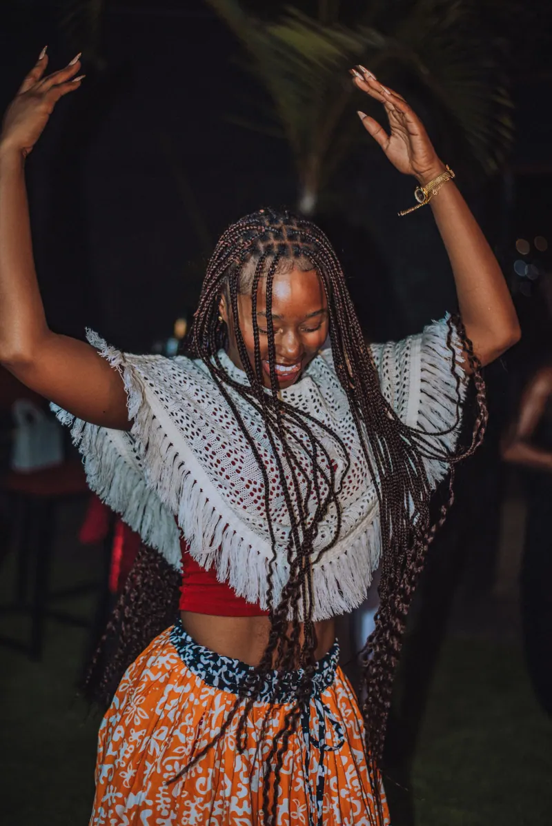 A woman in a white crochet top dances with both arms in the air, braids flying