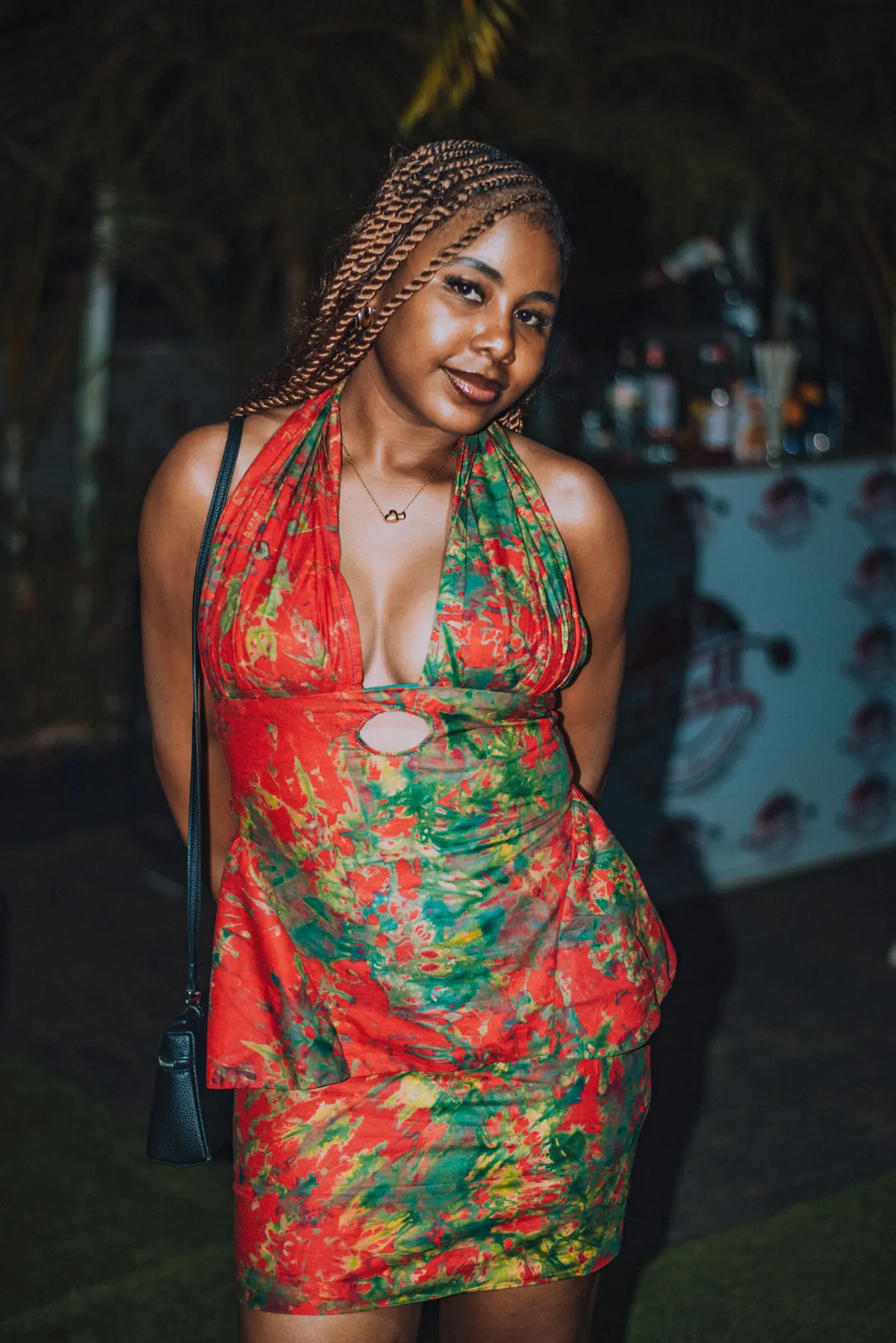 A woman with braids in a red and green floral halter dress stands with a bag on her shoulder