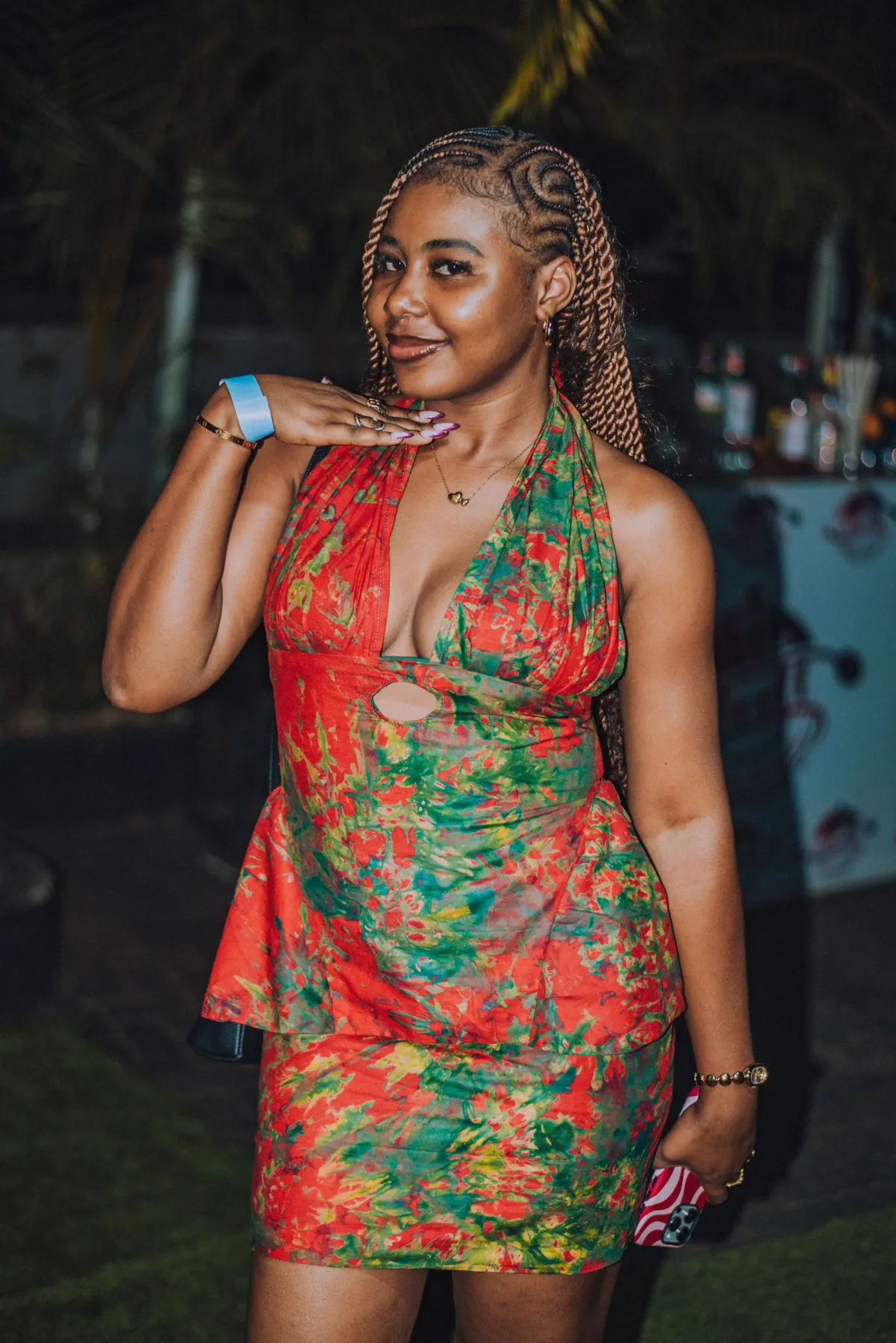 A woman in a red and green floral dress poses with a hand under her chin